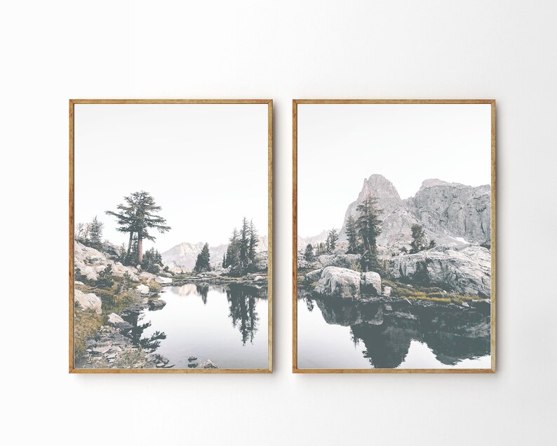 May include: Two framed landscape photographs of a mountain lake with a reflection of the surrounding mountains and trees. The lake is calm and still, and the water is a light blue color. The mountains are a dark gray color, and the trees are a light green color.