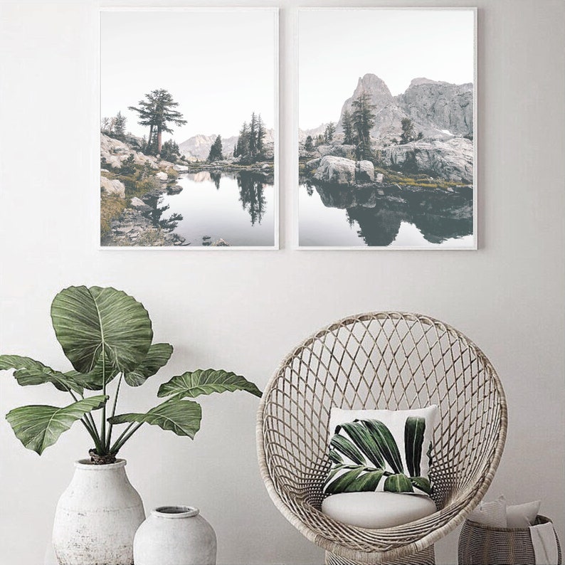 May include: Two framed landscape photographs of a lake surrounded by mountains and trees. The photos are hung on a white wall above a wicker chair with a green leaf patterned pillow.