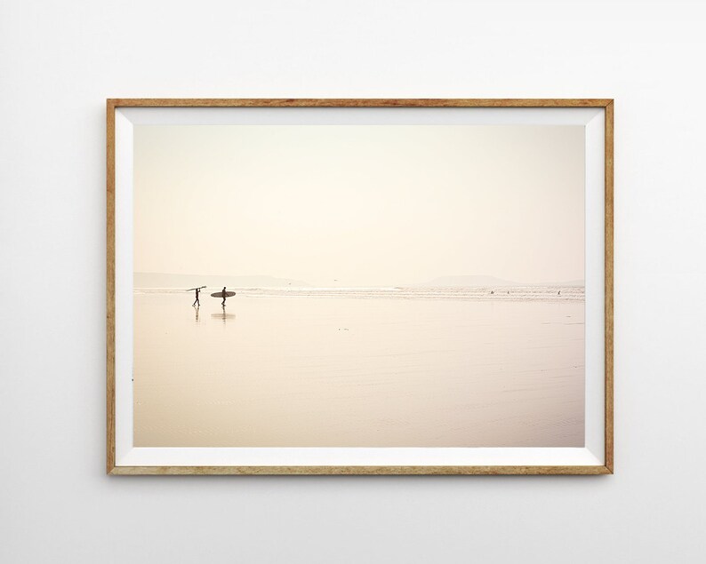May include: A framed photograph of two people walking on a sandy beach with surfboards. The beach is empty and the sky is a soft, pale pink.