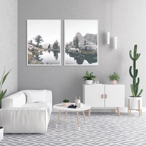 May include: A white couch with two pillows sits in a living room with a white cabinet, two plants, and two framed landscape photographs of a lake and mountains.