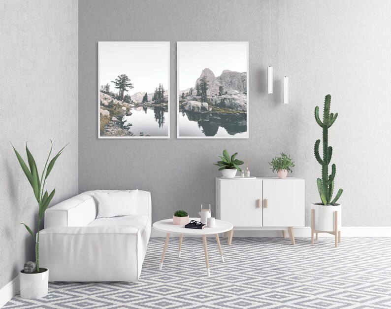 May include: A white couch with two pillows sits in a living room with a white cabinet, two plants, and two framed landscape photographs of a lake and mountains.