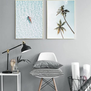May include: Two framed art prints. One shows a person floating in turquoise water, the other depicts two palm trees against a blue sky. The frames have gold borders. The art is displayed in a room with a lamp, chair, and cushions.