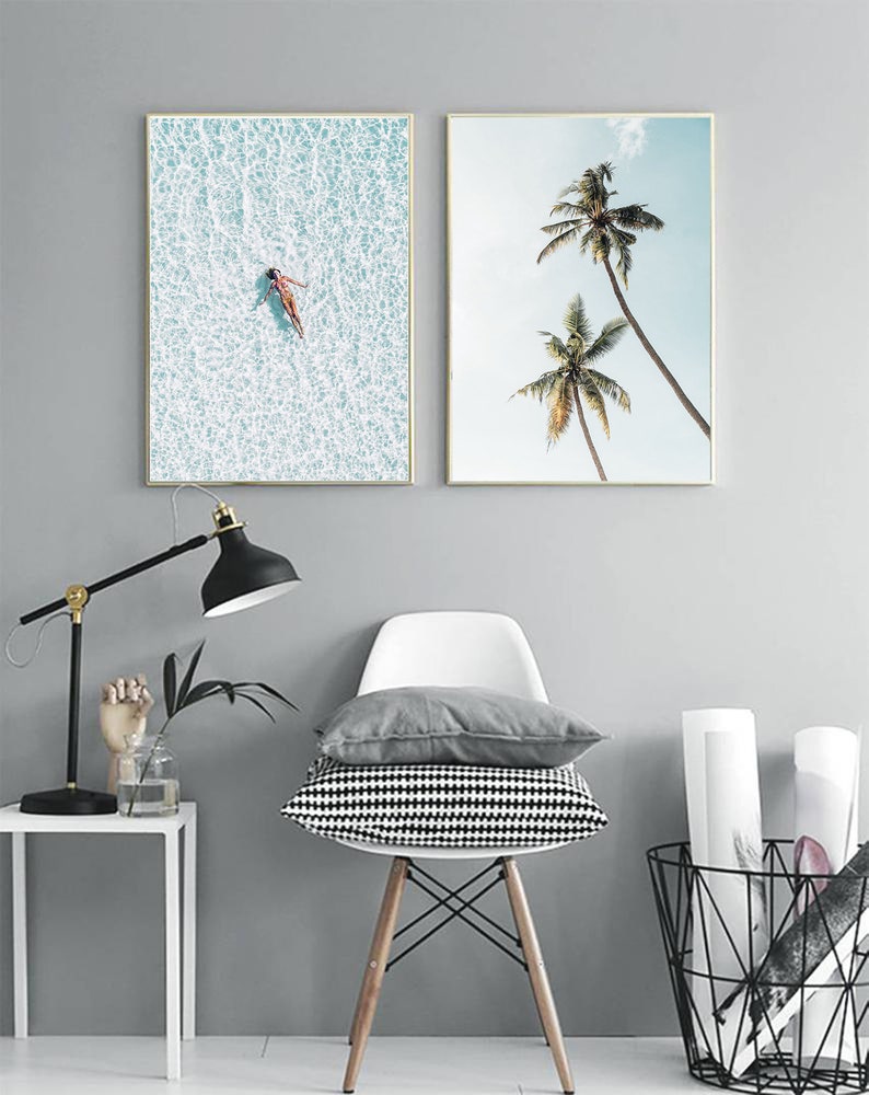 May include: Two framed art prints. One shows a person floating in turquoise water, the other depicts two palm trees against a blue sky. The frames have gold borders. The art is displayed in a room with a lamp, chair, and cushions.