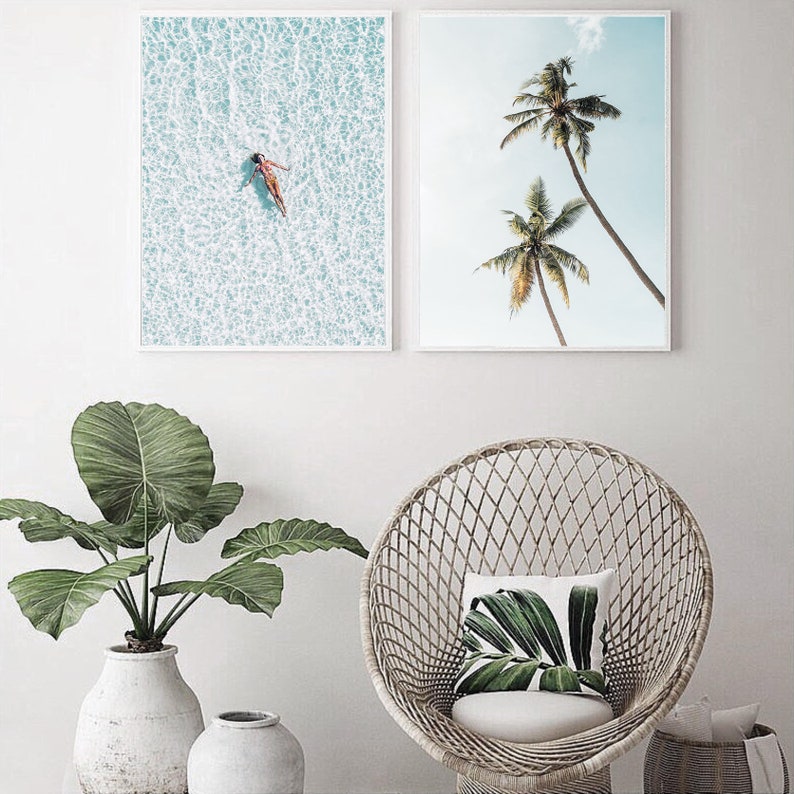 May include: Two framed art prints. One shows a person floating in turquoise water, the other depicts two palm trees against a blue sky. A large potted plant and a wicker chair with a cushion are in the foreground.