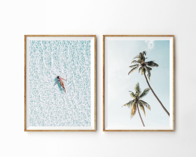 May include: Two framed art prints. One shows a person floating in clear turquoise water. The other depicts two tall palm trees against a light blue sky. Both prints are in wooden frames.
