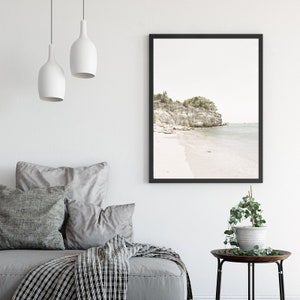 Puede incluir: Una fotograf&iacute;a enmarcada de una escena de playa con un acantilado rocoso y &aacute;rboles verdes en el fondo. La playa es de arena blanca y el agua es de color azul claro.