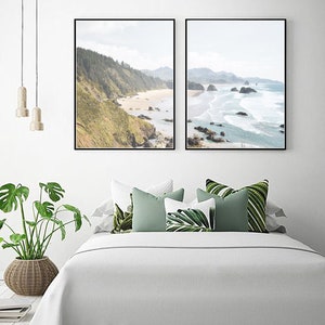 May include: Two framed landscape photographs of a beach with rocky cliffs and ocean waves. The photos are hung on a white wall above a white bed with green and white pillows.