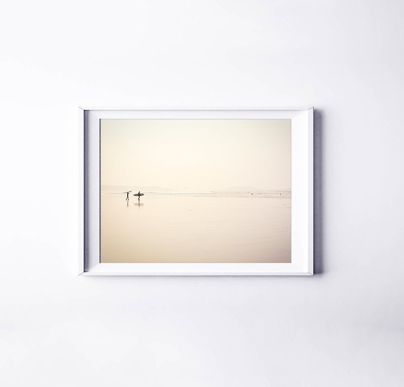 May include: A framed photograph of two people walking on a beach with surfboards. The beach is sandy and the water is calm and shallow. The sky is a pale yellow and the horizon is in the distance.