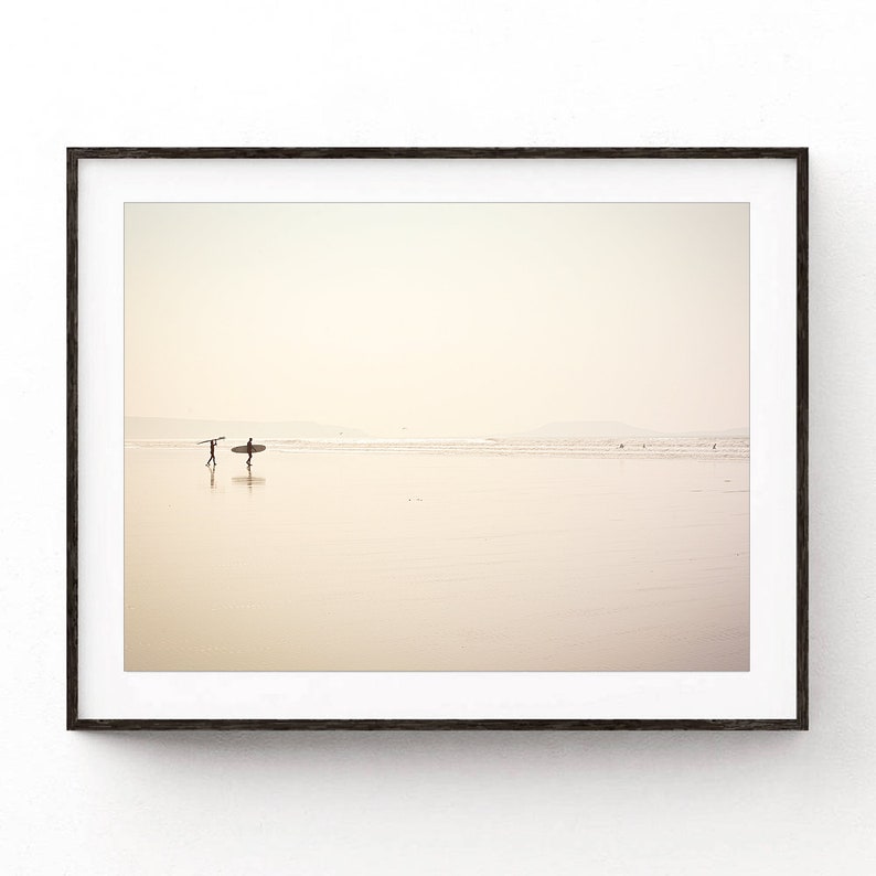 May include: A framed photograph of two people walking on a beach with a surfboard. The beach is empty and the sky is a pale yellow.