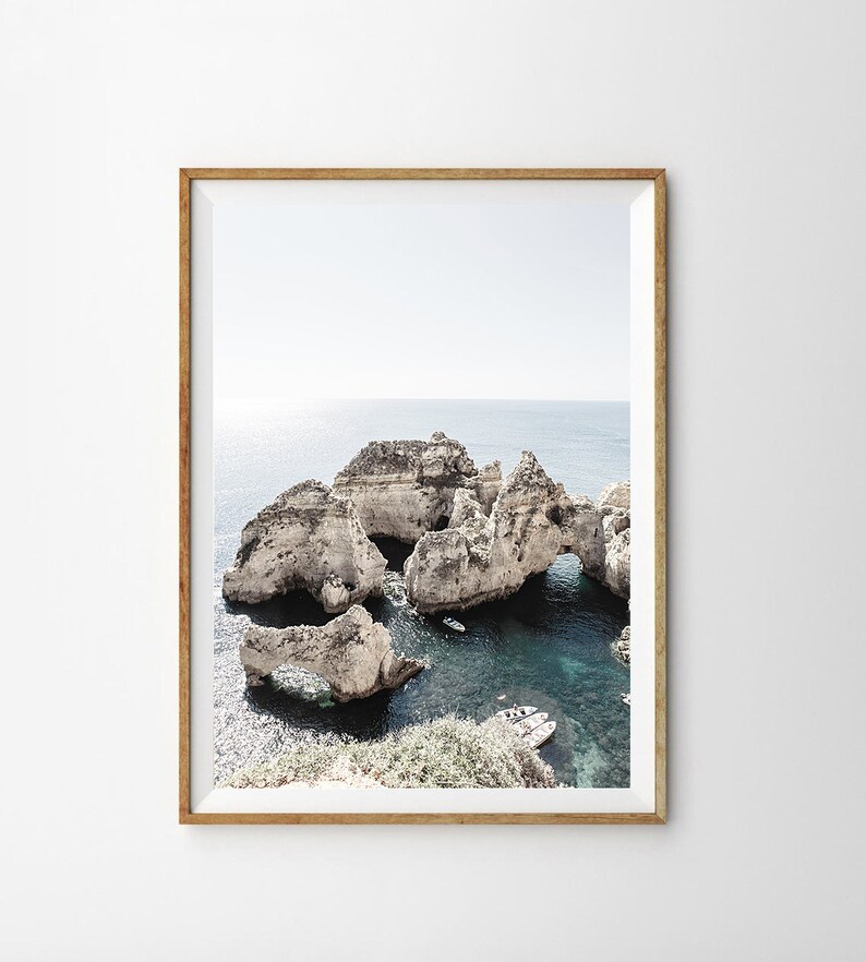 May include: A framed photograph of a rocky coastline with turquoise water and several small boats. The rocks are white and have many arches and caves. The sky is a light blue and the water is clear.