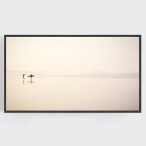 May include: A black framed photograph of a beach scene with two people walking on the sand with surfboards. The sky is a light beige color and the water is a light blue color.