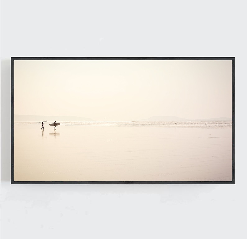 May include: A black framed photograph of a beach scene with two people walking on the sand with surfboards. The sky is a light beige color and the water is a light blue color.