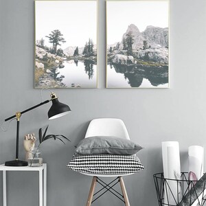 May include: Two framed landscape photographs of a mountain lake with a reflection of the mountains in the water. The lake is surrounded by trees and rocks. The photos are hung on a gray wall.