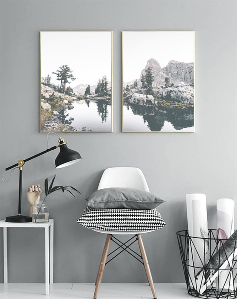 May include: Two framed landscape photographs of a mountain lake with a reflection of the mountains in the water. The lake is surrounded by trees and rocks. The photos are hung on a gray wall.