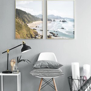 May include: Two framed landscape photographs of a beach with rocky cliffs and ocean waves. The photos are hung on a gray wall above a white chair with a black and white patterned cushion.