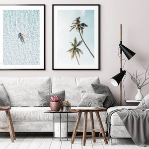 May include: Two framed art prints. One shows a person floating in turquoise water, the other depicts palm trees against a blue sky. The prints hang above a white sofa with grey cushions and wooden stools.