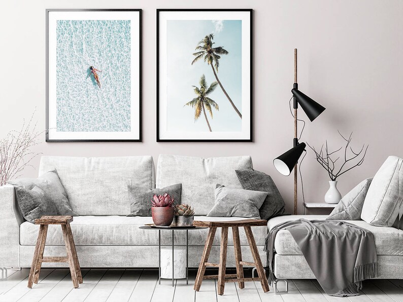 May include: Two framed art prints. One shows a person floating in turquoise water, the other depicts palm trees against a blue sky. The prints hang above a white sofa with grey cushions and wooden stools.