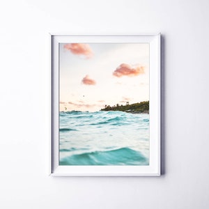 May include: A framed photograph of a turquoise ocean with waves crashing against the shore. The sky is a soft pink and orange with fluffy clouds. A small island with trees is visible in the distance.