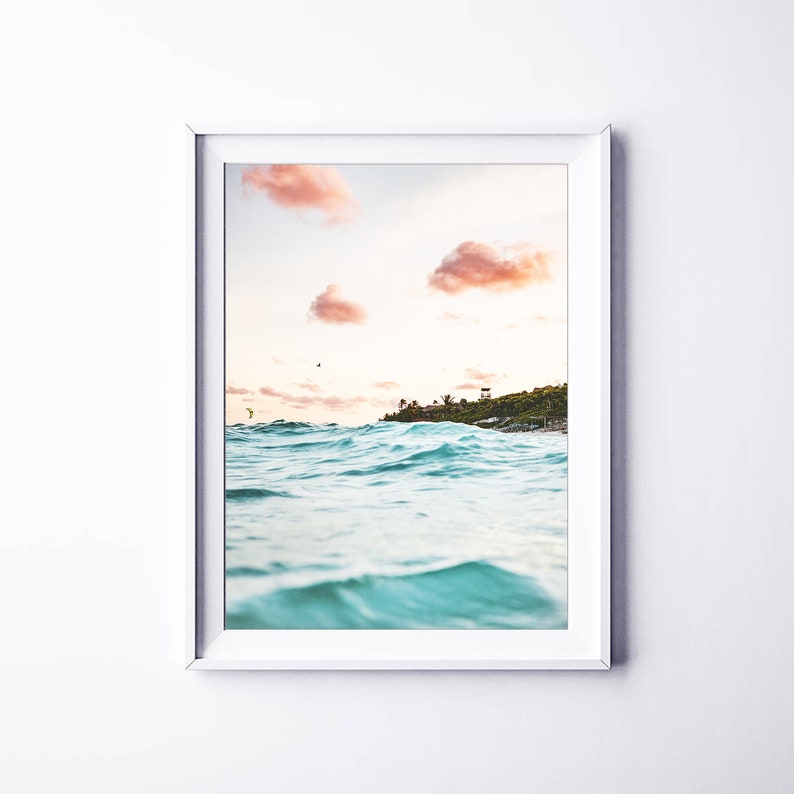 May include: A framed photograph of a turquoise ocean with waves crashing against the shore. The sky is a soft pink and orange with fluffy clouds. A small island with trees is visible in the distance.