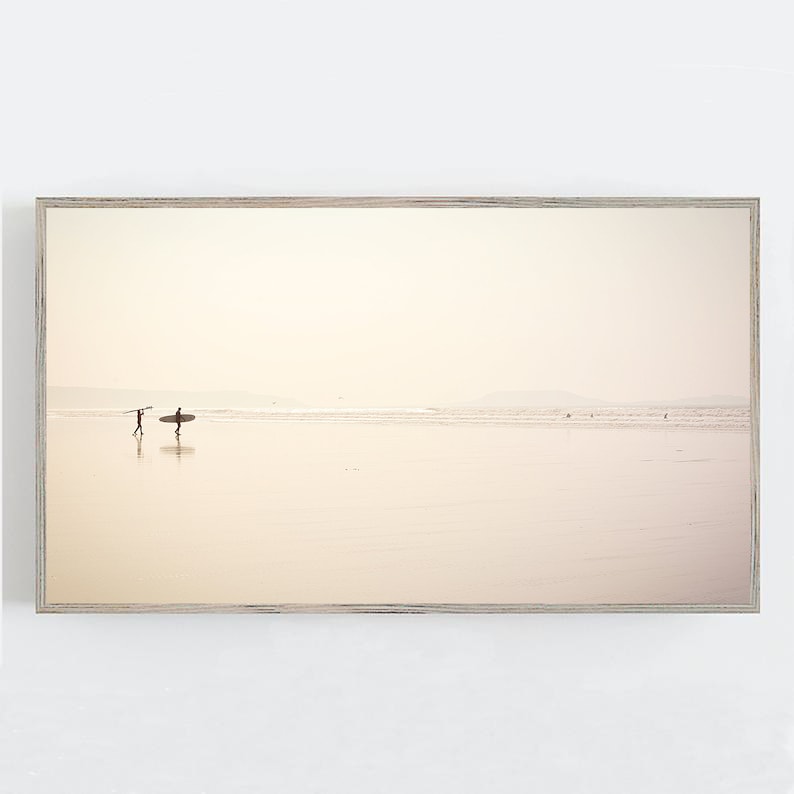 May include: A framed photograph of a beach scene with two people walking on the sand with surfboards. The sky is a soft, pale pink and the water is a light blue. The beach is empty except for the two people.