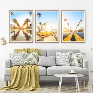 May include: Three framed photographs of a beach scene with palm trees, a bicycle, and a road with yellow lines. The photos are hung on a white wall above a gray couch with pillows and a throw blanket.