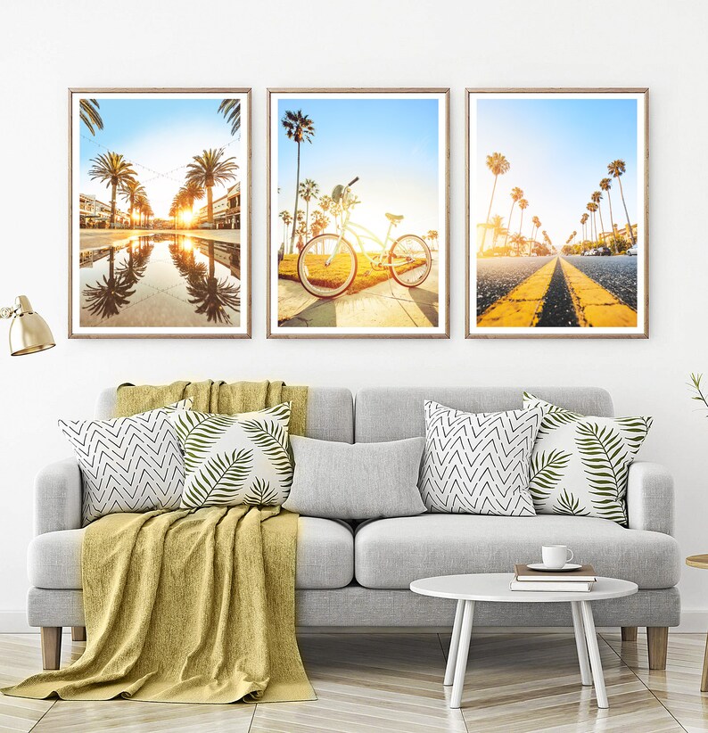 May include: Three framed photographs of a beach scene with palm trees, a bicycle, and a road with yellow lines. The photos are hung on a white wall above a gray couch with pillows and a throw blanket.