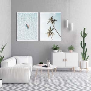 May include: Two framed art prints. One shows a person floating in turquoise water, the other depicts two palm trees against a blue sky. The prints are displayed in a modern living room setting with white furniture and plants.