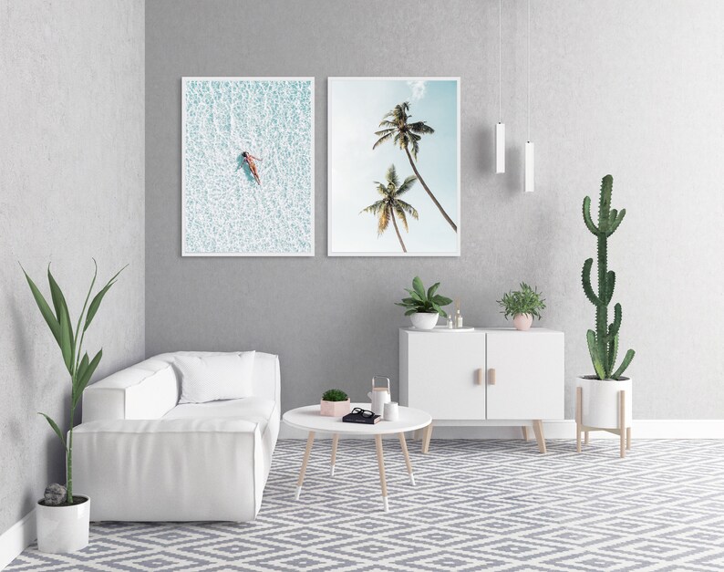 May include: Two framed art prints. One shows a person floating in turquoise water, the other depicts two palm trees against a blue sky. The prints are displayed in a modern living room setting with white furniture and plants.