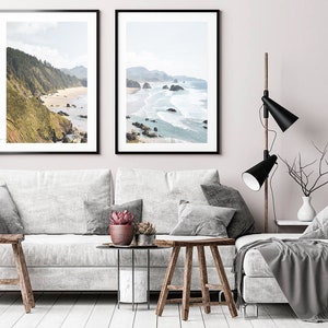 May include: Two framed landscape photographs of a rocky coastline with ocean waves crashing on the shore. The photos are hanging on a light pink wall above a white sofa with gray throw pillows and a gray throw blanket.