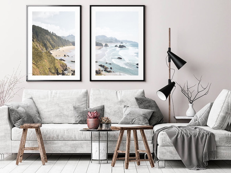 May include: Two framed landscape photographs of a rocky coastline with ocean waves crashing on the shore. The photos are hanging on a light pink wall above a white sofa with gray throw pillows and a gray throw blanket.