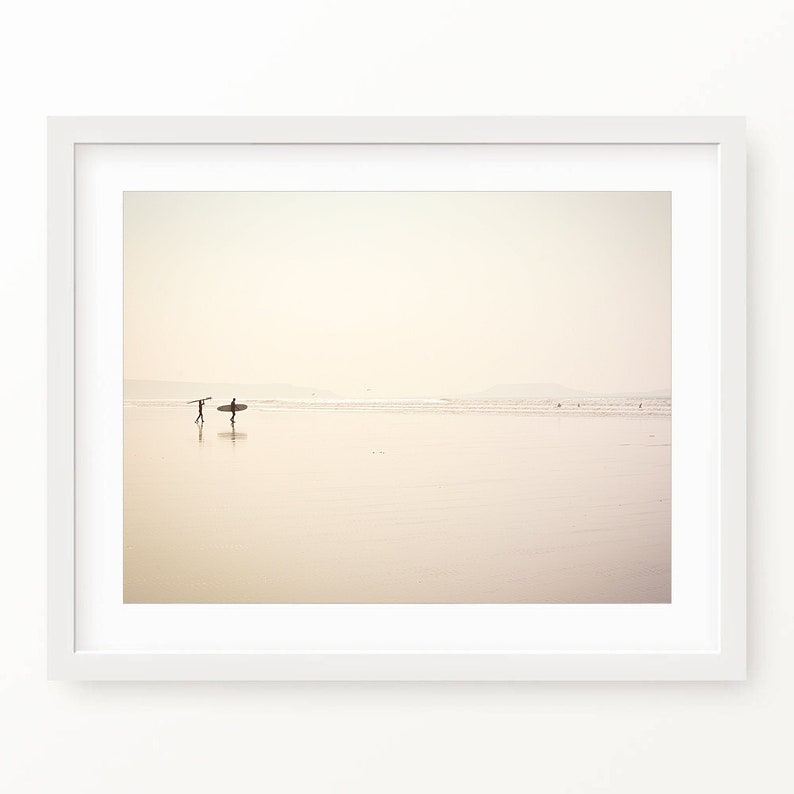 May include: Two people walk along a sandy beach carrying surfboards. The beach is empty except for a few people in the distance. The sky is a pale pink and the water is a light blue.