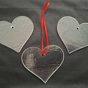 May include: Three clear heart-shaped decorations. One heart has a red ribbon for hanging. The hearts are made of a transparent material and are displayed on a black surface. Each heart has a small hole at the top.