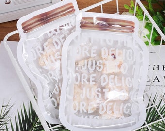 May include: Two clear plastic reusable food storage bags with a white zipper closure. The bags are shaped like mason jars and have the text "Just to the more delicious" printed on them.