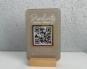 May include: A gray acrylic sign with a gold frame around a QR code. The sign reads "Purelivity Med Spa + Wellness" and "Scan the QR code for our services www.purelivitymedspa.com".