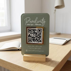 Op de afbeelding: Een groen rechthoekig bord met afgeronde hoeken, met de tekst "Purelivty Med Spa + Wellness" en een QR-code in een gouden lijst. Het bord is gemonteerd op een houten voet. De tekst "Scan the QR code for our services" is ook zichtbaar.
