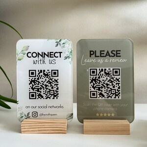 May include: Two acrylic signs with QR codes and text. The left sign is white with green foliage and says "CONNECT with us" and "on our social networks @bycraftqueen". The right sign is green and says "PLEASE Leave us a review" and "Scan the QR code with your phone camera". Both signs have a five-star rating.