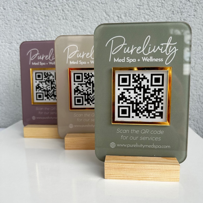 May include: Three acrylic signs with wooden bases, each featuring a different color and a gold-framed QR code. The signs advertise a med spa and wellness center called "Purelivity" with the website address "www.purelivitymedspa.com".