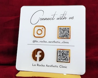May include: White sign with gold accents, featuring social media icons and QR codes for La Rocka Aesthetic Clinic. The text reads "Connect with us" and "@la_rocka_aesthetic_clinic".