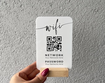 May include: A rectangular white acrylic sign with rounded corners, featuring the word wifi in black script, a black QR code, and the text NETWORK Your Network Here and PASSWORD Your Network Here, held in a light-colored wooden base stand.
