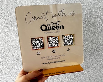 May include: A white sign with the text "Connect with us by Craft Queen" and a place for a logo. The sign has three gold QR codes with icons for Instagram, Facebook, and TikTok. The text "Scan the QR code with your phone camera" and the website address "www.ByCraftQueen.com" are also on the sign.