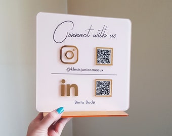 Op de afbeelding: Wit bord met gouden letters die "Connect with us" zeggen. Het bord heeft gouden pictogrammen voor Instagram, LinkedIn en twee QR-codes. De Instagram-handle is @klesisjunior.meaux en de LinkedIn-naam is Binta Badji.