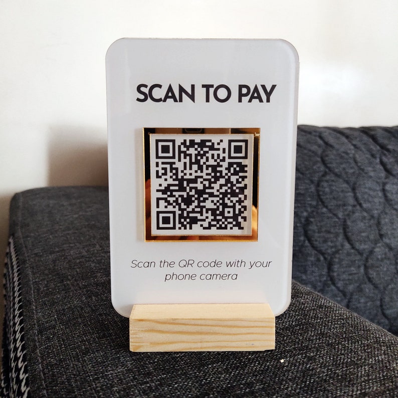 May include: White acrylic sign with the text "SCAN TO PAY" and a gold framed QR code. The sign is on a wooden stand. The text below the QR code reads "Scan the QR code with your phone camera."