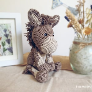 Puede incluir: Un juguete de burro de crochet marrón y blanco sentado sobre una superficie marrón claro. El burro tiene una cola y una crin largas y esponjosas.