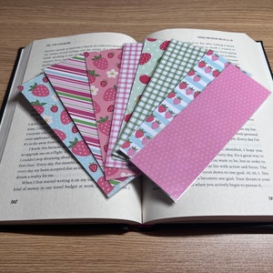 May include: A collection of colorful paper bookmarks with various patterns, including strawberries, stripes, and polka dots, fanned out over an open book. The bookmarks are in shades of pink, green, and blue.