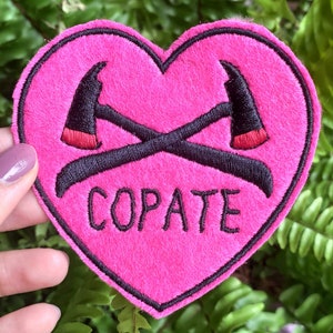 Puede incluir: Parche de fieltro rosa en forma de coraz&oacute;n con hachas cruzadas bordadas en negro y la palabra "COPATE" en negro.