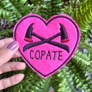Puede incluir: Un parche de fieltro rosa en forma de coraz&oacute;n con hachas cruzadas bordadas en negro y la palabra "COPATE" en letras negras.
