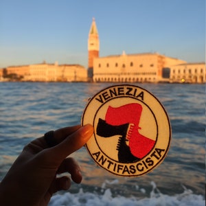 Può includere: Un cerotto circolare bianco con testo nero che recita "Venezia Antifascista". Il cerotto presenta un stivale rosso con un'ascia nera al suo interno. Il cerotto è tenuto davanti a uno sfondo sfocato di uno skyline cittadino con una torre alta.
