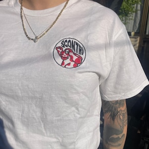 Peut inclure: T-shirt blanc avec un écusson brodé circulaire avec le mot "SCONTR" et une grenouille de dessin animé rouge et blanche. Le t-shirt est fait d'une matière douce.