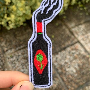 Può includere: Toppa ricamata nera e rossa di una bottiglia di salsa piccante con un logo verde e rosso. La bottiglia ha la forma di una mano con un pugno alzato.