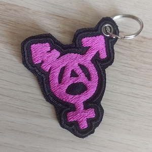 Può includere: Un portachiavi in tessuto nero con un simbolo transgender rosa e un simbolo anarchico al suo interno.